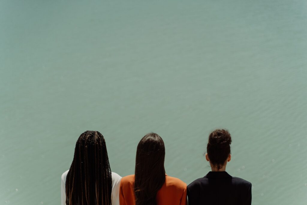 Three women seen from behind, gazing at a calm lake under clear daylight.